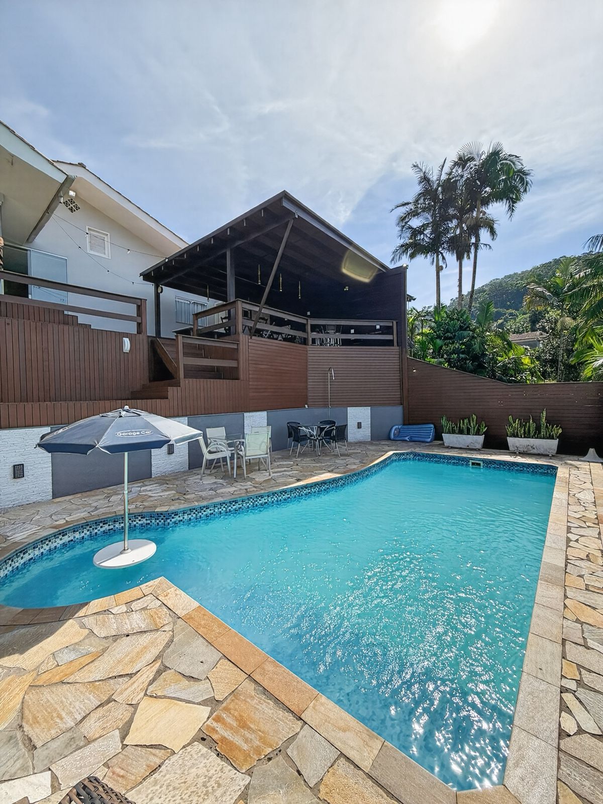 Piscina com piso de pedra e ombrelone, com o deck coberto da área de festas e a casa ao fundo